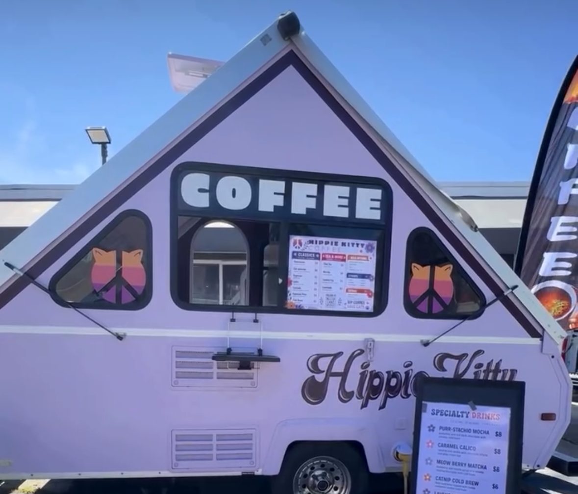 The Hippie Kitty Coffee lavender A-frame camper, serving coffee from its window with the specialty drinks board out front